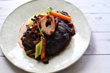 Beyaz bir tabakta renkli sebzelerle servis edilen kemikli kızarmış sığır eti. Tabakta yumuşak, fırında kızartılmış et ve mevsimlik sebzeler yer alıyor ve temiz ve minimalist bir şekilde sunuluyor.