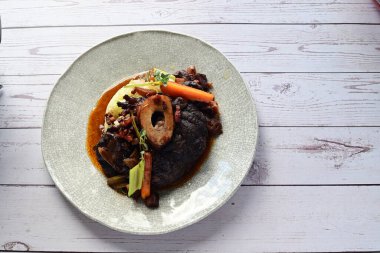 Beyaz bir tabakta renkli sebzelerle servis edilen kemikli kızarmış sığır eti. Tabakta yumuşak, fırında kızartılmış et ve mevsimlik sebzeler yer alıyor ve temiz ve minimalist bir şekilde sunuluyor.