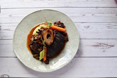 Beyaz bir tabakta renkli sebzelerle servis edilen kemikli kızarmış sığır eti. Tabakta yumuşak, fırında kızartılmış et ve mevsimlik sebzeler yer alıyor ve temiz ve minimalist bir şekilde sunuluyor.