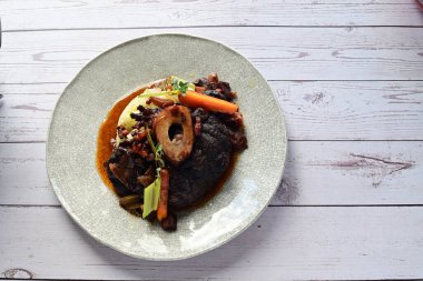 Beyaz bir tabakta renkli sebzelerle servis edilen kemikli kızarmış sığır eti. Tabakta yumuşak, fırında kızartılmış et ve mevsimlik sebzeler yer alıyor ve temiz ve minimalist bir şekilde sunuluyor.