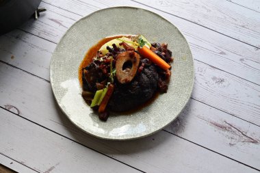 Beyaz bir tabakta renkli sebzelerle servis edilen kemikli kızarmış sığır eti. Tabakta yumuşak, fırında kızartılmış et ve mevsimlik sebzeler yer alıyor ve temiz ve minimalist bir şekilde sunuluyor.