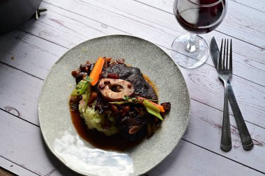 Sebzelerle servis edilen ve taze otlarla süslenen sığır eti osso buco 'sunun yakın çekimi. Kemik iliğiyle pişirilmiş yumuşak et, bu geleneksel İtalyan yemeğinin dokusunu ve derinliğini vurguluyor. 