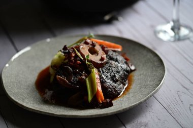Sebzelerle servis edilen ve taze otlarla süslenen sığır eti osso buco 'sunun yakın çekimi. Kemik iliğiyle pişirilmiş yumuşak et, bu geleneksel İtalyan yemeğinin dokusunu ve derinliğini vurguluyor. 