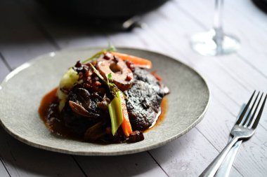 Sebzelerle servis edilen ve taze otlarla süslenen sığır eti osso buco 'sunun yakın çekimi. Kemik iliğiyle pişirilmiş yumuşak et, bu geleneksel İtalyan yemeğinin dokusunu ve derinliğini vurguluyor. 