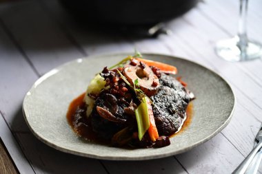Sebzelerle servis edilen ve taze otlarla süslenen sığır eti osso buco 'sunun yakın çekimi. Kemik iliğiyle pişirilmiş yumuşak et, bu geleneksel İtalyan yemeğinin dokusunu ve derinliğini vurguluyor. 