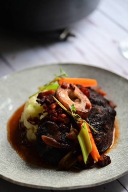 Sebzelerle servis edilen ve taze otlarla süslenen sığır eti osso buco 'sunun yakın çekimi. Kemik iliğiyle pişirilmiş yumuşak et, bu geleneksel İtalyan yemeğinin dokusunu ve derinliğini vurguluyor. 