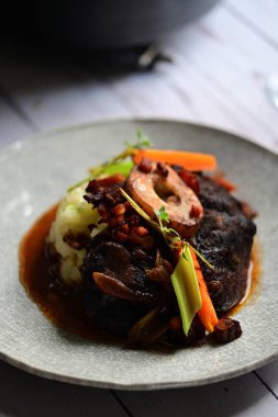 Yanında taze patates, kök sebze ve taze otlarla birlikte kırmızı şarap soslu kemikli haşlanmış sığır eti servis edilir..