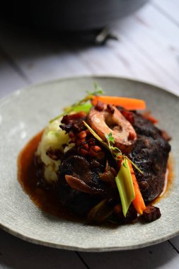 Yanında taze patates, kök sebze ve taze otlarla birlikte kırmızı şarap soslu kemikli haşlanmış sığır eti servis edilir..