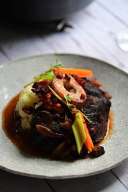 Yanında taze patates, kök sebze ve taze otlarla birlikte kırmızı şarap soslu kemikli haşlanmış sığır eti servis edilir..