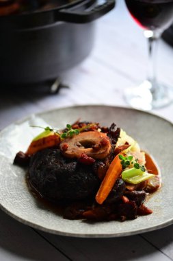 Yanında taze patates, kök sebze ve taze otlarla birlikte kırmızı şarap soslu kemikli haşlanmış sığır eti servis edilir..