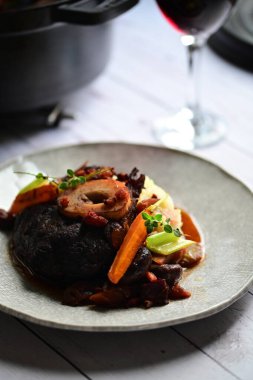 Yanında taze patates, kök sebze ve taze otlarla birlikte kırmızı şarap soslu kemikli haşlanmış sığır eti servis edilir..
