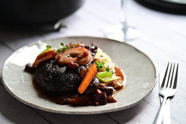 Yanında taze patates, kök sebze ve taze otlarla birlikte kırmızı şarap soslu kemikli haşlanmış sığır eti servis edilir..