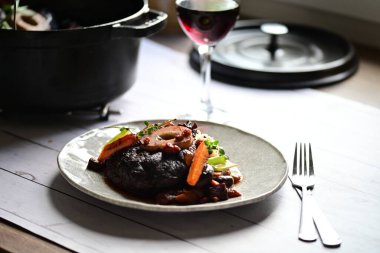 Yanında taze patates, kök sebze ve taze otlarla birlikte kırmızı şarap soslu kemikli haşlanmış sığır eti servis edilir..