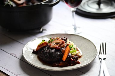 Yanında taze patates, kök sebze ve taze otlarla birlikte kırmızı şarap soslu kemikli haşlanmış sığır eti servis edilir..