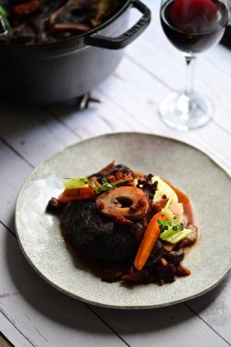 Yanında taze patates, kök sebze ve taze otlarla birlikte kırmızı şarap soslu kemikli haşlanmış sığır eti servis edilir..