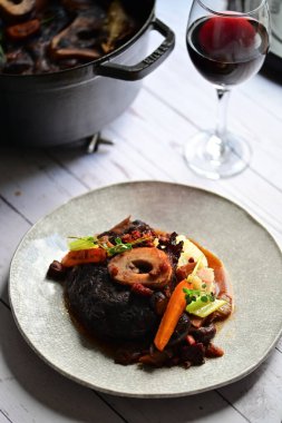 Yanında taze patates, kök sebze ve taze otlarla birlikte kırmızı şarap soslu kemikli haşlanmış sığır eti servis edilir..