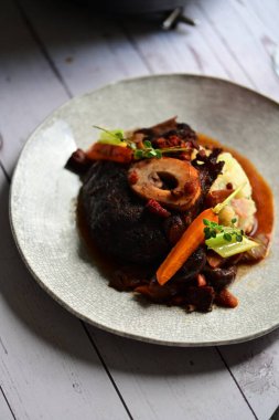 Yanında taze patates, kök sebze ve taze otlarla birlikte kırmızı şarap soslu kemikli haşlanmış sığır eti servis edilir..