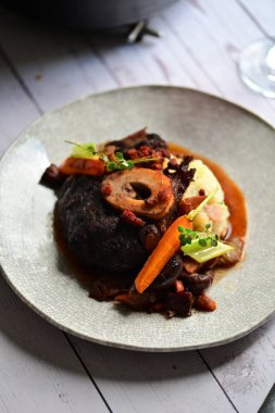 Yanında taze patates, kök sebze ve taze otlarla birlikte kırmızı şarap soslu kemikli haşlanmış sığır eti servis edilir..