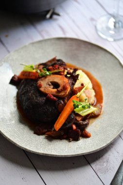 Yanında taze patates, kök sebze ve taze otlarla birlikte kırmızı şarap soslu kemikli haşlanmış sığır eti servis edilir..