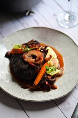 Yanında taze patates, kök sebze ve taze otlarla birlikte kırmızı şarap soslu kemikli haşlanmış sığır eti servis edilir..