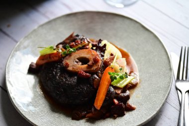 Yanında taze patates, kök sebze ve taze otlarla birlikte kırmızı şarap soslu kemikli haşlanmış sığır eti servis edilir..