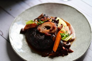 Yanında taze patates, kök sebze ve taze otlarla birlikte kırmızı şarap soslu kemikli haşlanmış sığır eti servis edilir..