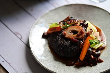 Yanında taze patates, kök sebze ve taze otlarla birlikte kırmızı şarap soslu kemikli haşlanmış sığır eti servis edilir..