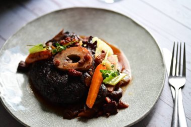 Yanında taze patates, kök sebze ve taze otlarla birlikte kırmızı şarap soslu kemikli haşlanmış sığır eti servis edilir..