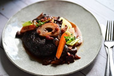 Yanında taze patates, kök sebze ve taze otlarla birlikte kırmızı şarap soslu kemikli haşlanmış sığır eti servis edilir..