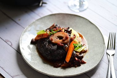 Yanında taze patates, kök sebze ve taze otlarla birlikte kırmızı şarap soslu kemikli haşlanmış sığır eti servis edilir..