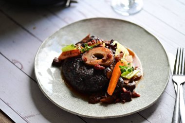 Yanında taze patates, kök sebze ve taze otlarla birlikte kırmızı şarap soslu kemikli haşlanmış sığır eti servis edilir..