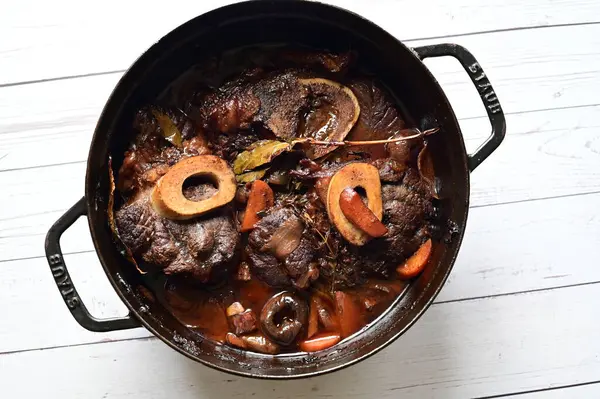 Kırmızı şarapta pişirilmiş kemikli yumuşak sığır eti, taze otlar ve tek bir kırsal siyah tencerede kök sebzelerle servis edilir..