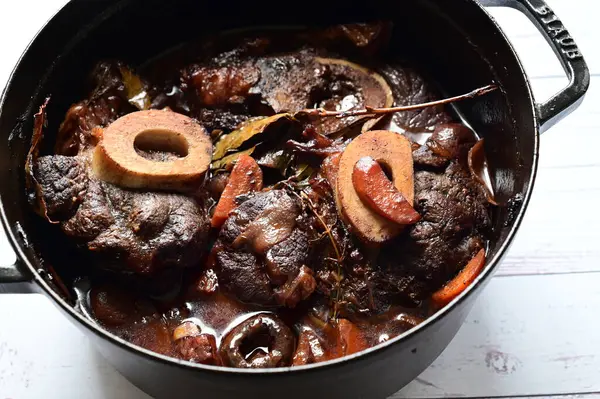 Kırmızı şarapta pişirilmiş kemikli yumuşak sığır eti, taze otlar ve tek bir kırsal siyah tencerede kök sebzelerle servis edilir..