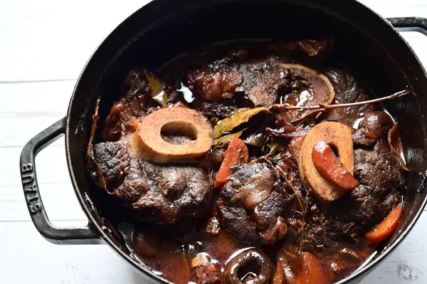 Kırmızı şarapta pişirilmiş kemikli yumuşak sığır eti, taze otlar ve tek bir kırsal siyah tencerede kök sebzelerle servis edilir..