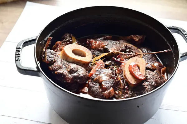 Kırmızı şarapta pişirilmiş kemikli yumuşak sığır eti, taze otlar ve tek bir kırsal siyah tencerede kök sebzelerle servis edilir..