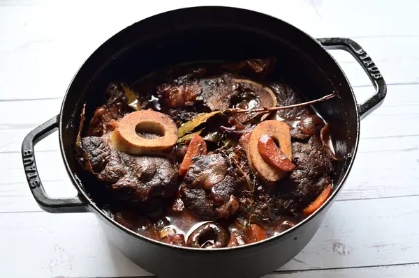 Kırmızı şarapta pişirilmiş kemikli yumuşak sığır eti, taze otlar ve tek bir kırsal siyah tencerede kök sebzelerle servis edilir..