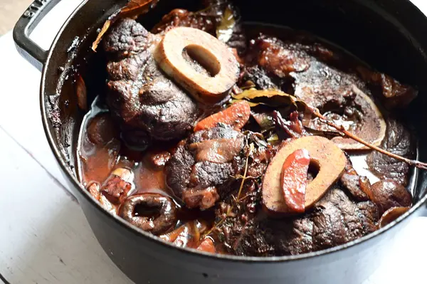 Kırmızı şarapta pişirilmiş kemikli yumuşak sığır eti, taze otlar ve tek bir kırsal siyah tencerede kök sebzelerle servis edilir..