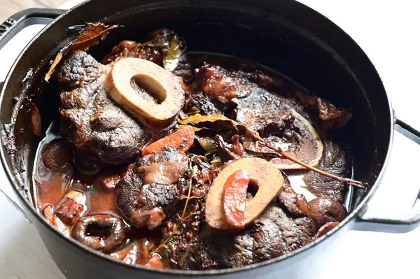 Kırmızı şarapta pişirilmiş kemikli yumuşak sığır eti, taze otlar ve tek bir kırsal siyah tencerede kök sebzelerle servis edilir..