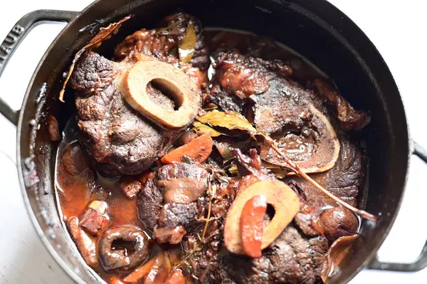 Kırmızı şarapta pişirilmiş kemikli yumuşak sığır eti, taze otlar ve tek bir kırsal siyah tencerede kök sebzelerle servis edilir..