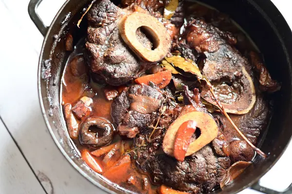 Kırmızı şarapta pişirilmiş kemikli yumuşak sığır eti, taze otlar ve tek bir kırsal siyah tencerede kök sebzelerle servis edilir..