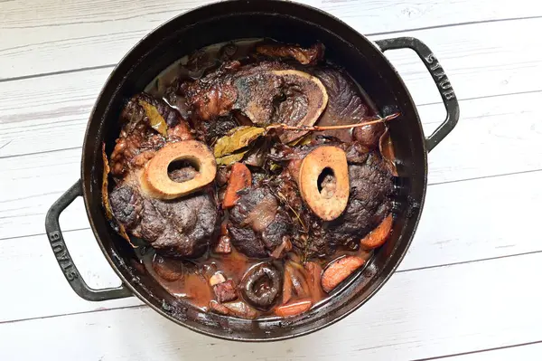 Kırmızı şarapta pişirilmiş kemikli yumuşak sığır eti, taze otlar ve tek bir kırsal siyah tencerede kök sebzelerle servis edilir..