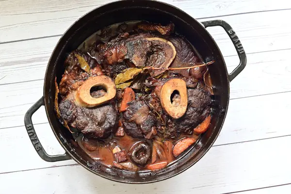 Kırmızı şarapta pişirilmiş kemikli yumuşak sığır eti, taze otlar ve tek bir kırsal siyah tencerede kök sebzelerle servis edilir..