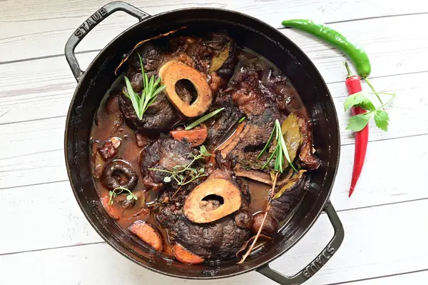 Kırmızı şarapta pişirilmiş kemikli yumuşak sığır eti, taze otlar ve tek bir kırsal siyah tencerede kök sebzelerle servis edilir..