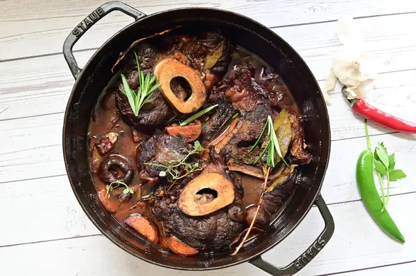 Kırmızı şarapta pişirilmiş kemikli yumuşak sığır eti, taze otlar ve tek bir kırsal siyah tencerede kök sebzelerle servis edilir..