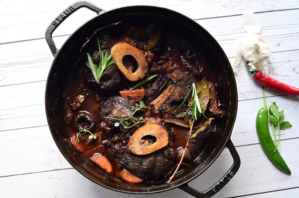 Kırmızı şarapta pişirilmiş kemikli yumuşak sığır eti, taze otlar ve tek bir kırsal siyah tencerede kök sebzelerle servis edilir..