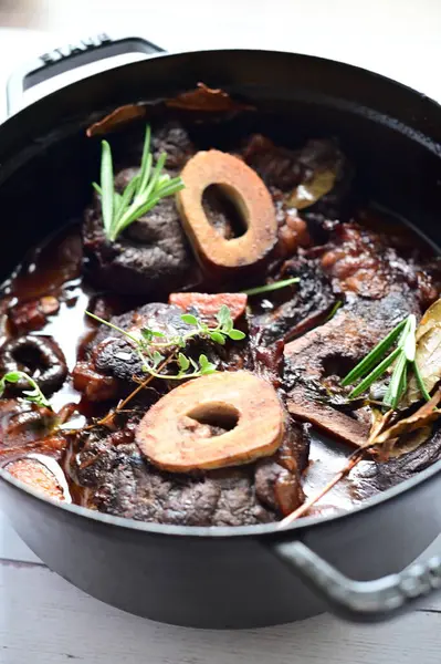 Kırmızı şarapta pişirilmiş kemikli yumuşak sığır eti, taze otlar ve tek bir kırsal siyah tencerede kök sebzelerle servis edilir..