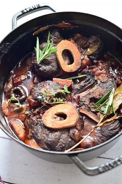 Kırmızı şarapta pişirilmiş kemikli yumuşak sığır eti, taze otlar ve tek bir kırsal siyah tencerede kök sebzelerle servis edilir..