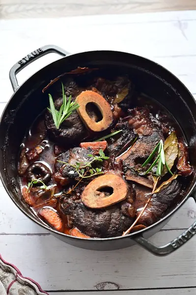Yanında taze patates, kök sebze ve taze otlarla birlikte kırmızı şarap soslu kemikli haşlanmış sığır eti servis edilir. Beyaz bir tabakta parlak bir arkaplanda sunulan bu çanak, klasik konforlu yiyecekleri inceliyor.