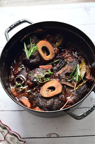 Yanında taze patates, kök sebze ve taze otlarla birlikte kırmızı şarap soslu kemikli haşlanmış sığır eti servis edilir. Beyaz bir tabakta parlak bir arkaplanda sunulan bu çanak, klasik konforlu yiyecekleri inceliyor.