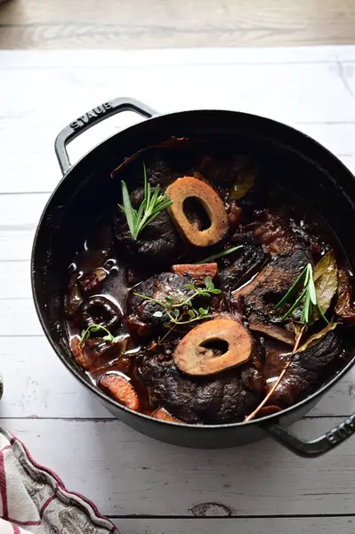 Yanında taze patates, kök sebze ve taze otlarla birlikte kırmızı şarap soslu kemikli haşlanmış sığır eti servis edilir. Beyaz bir tabakta parlak bir arkaplanda sunulan bu çanak, klasik konforlu yiyecekleri inceliyor.